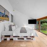 Schlafzimmer mit Bett auf Paletten, Fernseher und großen Fenstern zum Garten.