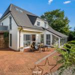 Haus mit Terrasse, Garten und Sicht auf Bäume unter blauem Himmel.
