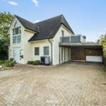 Haus mit Garage, Balkon und gepflastertem Hof. Umgeben von Grün und einem Holzzaun.