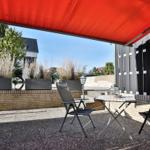 Terrasse mit rotem Sonnensegel, Tisch und zwei Stühlen. Hintergrund: Garten und Nachbarhaus.