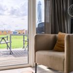 Sitzgelegenheit mit Kissen und Ständerventilator vor Glasfront mit Blick auf Terrasse und Grünfläche.