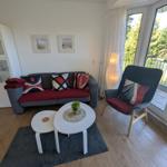 Wohnzimmer mit Sofa, Sessel und Tisch. Durchgang zur Terrasse mit Blick auf Grünflächen.