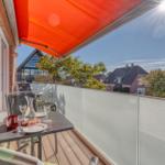 Terrasse mit Tisch, Stühlen und orangefarbenem Sonnenschutz. Blick auf Wohnhäuser und Bäume.