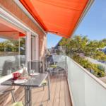 Terrasse mit Tisch, Stühlen und orangefarbenem Markisenüberdachung. Blick auf Straße und Bäume.