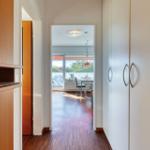 Flur mit Holzboden führt in Essbereich mit Tisch und Stühlen. Links Holzregal, rechts Schrank.