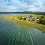 Häuser am Ufer eines Sees mit Grünland und Wald im Hintergrund.