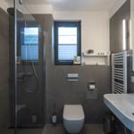 Badezimmer mit Dusche, Toilette und Waschbecken. Fenster mit Jalousie und Heizkörper.