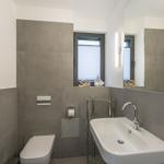 Badezimmer mit Waschbecken, Toilette und Fenster mit Jalousie.