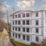 Weißes Mehrfamilienhaus mit blauem Schild und Fenstern. Umgeben von Bäumen und Grünflächen.