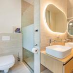 Badezimmer mit Dusche, Toilette und zwei Waschbecken mit beleuchteten Spiegeln.