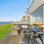 Terrasse mit Sofa, Tisch und Grill. Blick auf Wasser und Grünfläche.