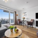 Wohnbereich mit Balkon, TV und Esstisch. Holzboden und große Fenster.