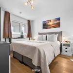 Schlafzimmer mit Bett, Nachttisch und Wandbild. Holzboden und Fenster mit Vorhängen.