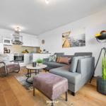 Wohnbereich mit Sofa, Sessel und Küchenzeile. Wandbild und Stehlampe sichtbar.