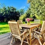 Holztisch mit Stühlen auf Terrasse, Blumen und Grill im Hintergrund.