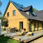 Haus mit Holzterrasse, Sonnenkollektoren und Gartenmöbel unter blauem Himmel.