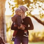 Eine Frau hält ein Kind im Freien. Ein Hund steht daneben. Bäume und Sonnenlicht sind sichtbar.