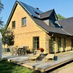 Haus mit Holzterrasse, Sonnenkollektoren und Gartenmöbel unter blauem Himmel.