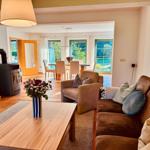 Wohnzimmer mit Sofa, Sessel und Holztisch. Im Hintergrund Essbereich mit Blick auf Garten.