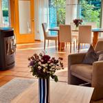 Wohn- und Essbereich mit Holztisch, Sofa und Kaminofen. Große Fenster mit Blick auf Garten.