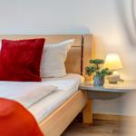 Hölzernes Bett mit weißen Kissen und rotem Kissen. Nachttisch mit Lampe und Pflanze.