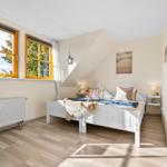 Schlafzimmer mit weißem Bett, Holzboden und zwei Fenstern mit orangefarbenen Rahmen.