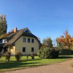 Ein gelbes Haus mit Strohdach, umgeben von Grün und Bäumen unter blauem Himmel.