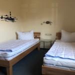 Zwei Einzelbetten mit Matratzen und Kissen. Wanddekoration mit Leuchtturm. Zwischen den Betten steht ein kleiner Tisch.