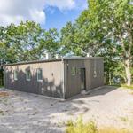 Moderne Holzhütte mit Fenstern und Tür, umgeben von Bäumen und Kies.