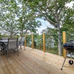 Holzterrasse mit Grill, Tisch und Stühlen unter Bäumen.