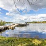 Boot liegt am Steg eines ruhigen Sees mit Gras und Bäumen im Hintergrund.