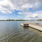 Holzsteg mit Boot liegt am Wasser. Im Hintergrund sind Bäume und ein Turm sichtbar.
