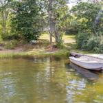 Ein kleines Boot liegt an einem Holzsteg am Ufer eines ruhigen Gewässers, umgeben von Grünflächen und Bäumen.