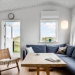 Wohnzimmer mit Sofa, Holztisch und Sitzgelegenheit. Fenster und Tür zeigen Landschaft.