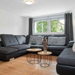 Ein Wohnzimmer mit grauem Sofa, Holzboden und Fenster mit Blick auf Grün.