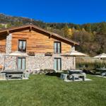 Haus mit Stein- und Holzfassade, Garten mit Tischen und Sonnenschirmen, Berglandschaft im Hintergrund.