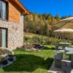 Holzhaus mit Steinmauer, Gartenbereich mit Tisch, Sonnenschirm und Blick auf Wald.