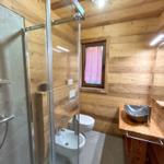 Badezimmer mit Dusche, Waschbecken und Toilette. Holzwandverkleidung und Fenster mit rotem Vorhang.