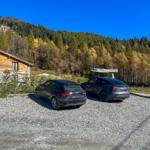 Zwei Autos stehen auf Kies vor einem Holzhaus mit Steinbasis.