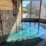 Innenpool mit Steinwand und Holzwand. Durch Fenster Blick auf Berglandschaft.