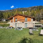 Haus mit Stein- und Holzfassade, Garten mit Liegestühlen und Sonnenschirmen, Berglandschaft im Hintergrund.