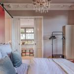 Schlafzimmer mit Bett, roten Vorhängen und Kronleuchter. Tür führt in Wohnbereich.