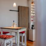 Esstisch mit Stühlen und einer Weinflasche. Hintergrund: Holzpaneel und Regal mit Geschirr.