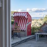 Terrasse mit Strandkorb, Tisch und Stuhl, Blick auf Wasser und Bäume.