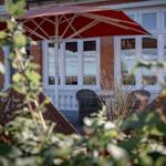 Terrasse mit rotem Sonnenschirm und zwei Stühlen vor einem Haus mit weißen Fenstern.