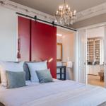 Schlafzimmer mit Bett, roter Schiebetür und Kronleuchter. Blick in die Küche und Bad.