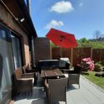 Terrasse mit Tisch, Stühlen und rotem Sonnenschirm. Grasfläche und Blumen im Hintergrund.