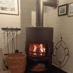 Kaminofen mit brennendem Feuer, Kaminrohr, Korb und Kerzenhalter an weißer Ziegelwand.