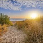 Sandweg führt durch Gras zu Strand und Meer bei Sonnenuntergang.