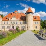 Schloss mit rotem Dach und grüner Umgebung. Pfad mit Bänken führt zum Eingang.